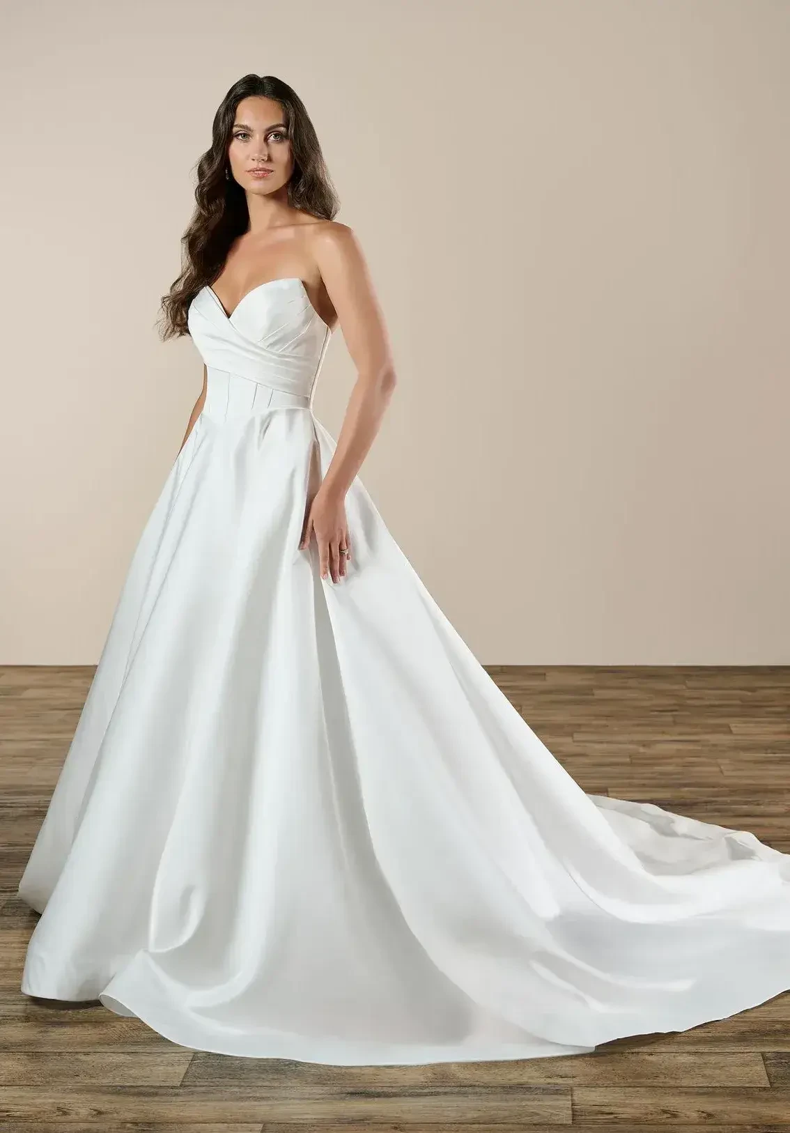 A woman in a strapless white wedding gown stands elegantly, with a flowing train and a neutral background.