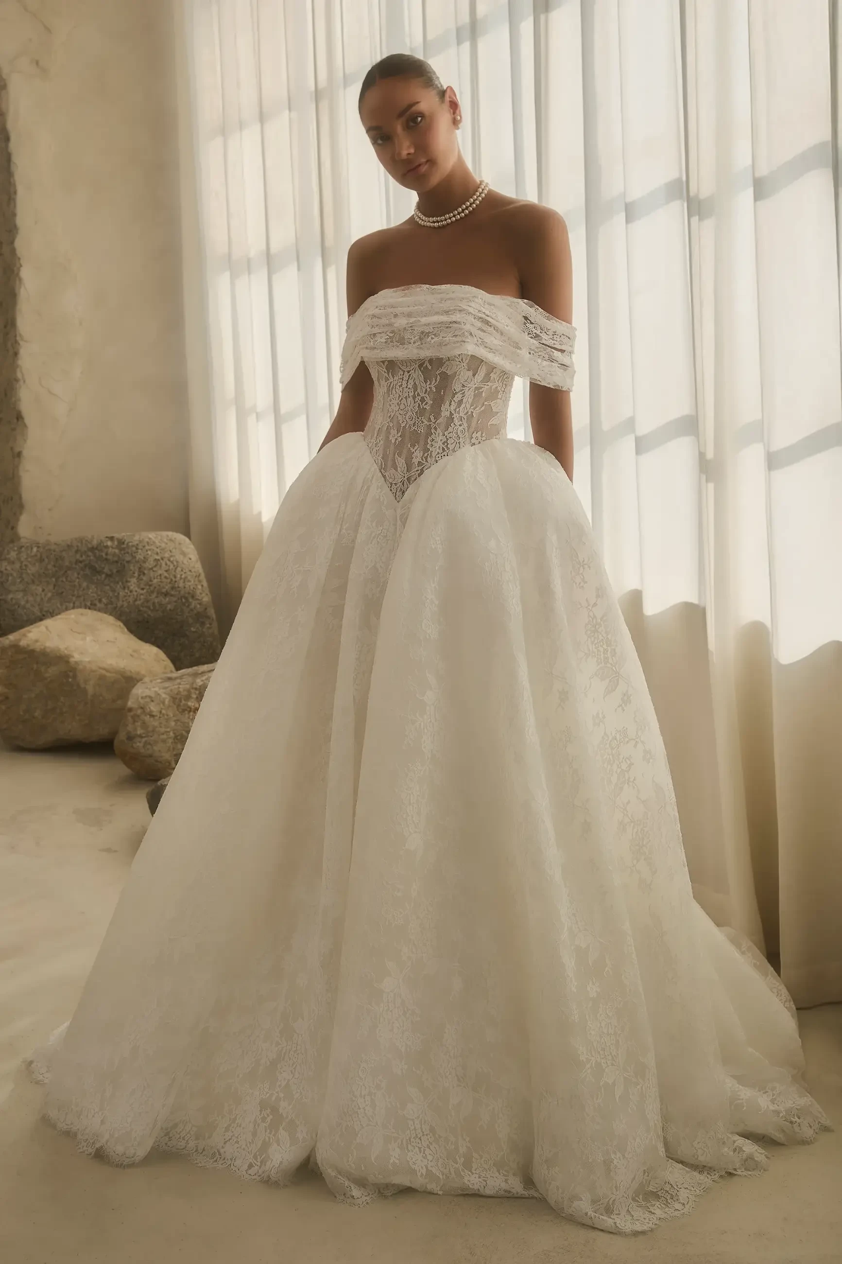 A woman in an elegant, off-shoulder lace wedding gown stands gracefully in soft natural light. The tone is serene and sophisticated, with stones in the background.