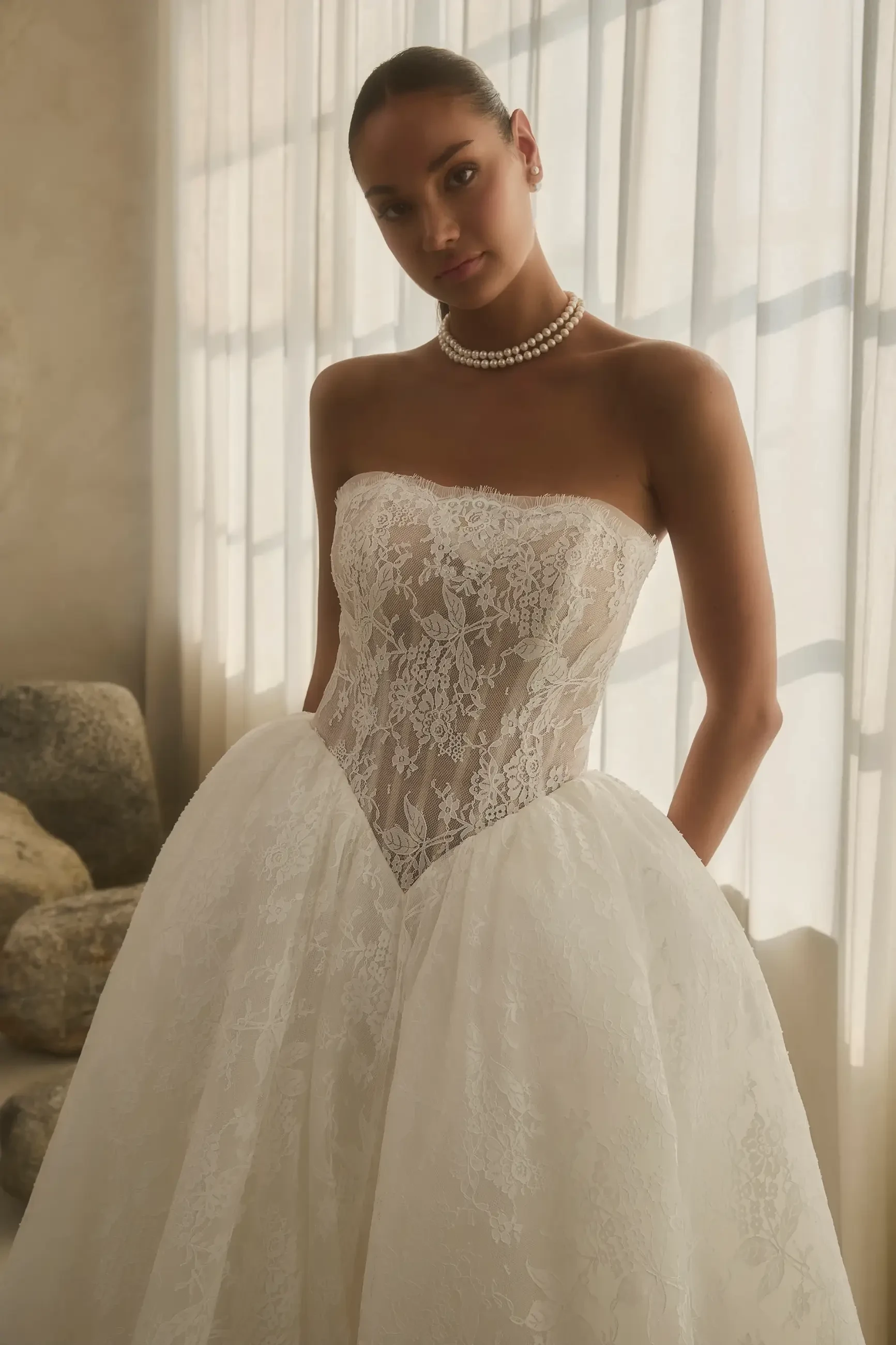 A model wearing a strapless wedding dress with lace detailing, posed in front of a light-filled window.