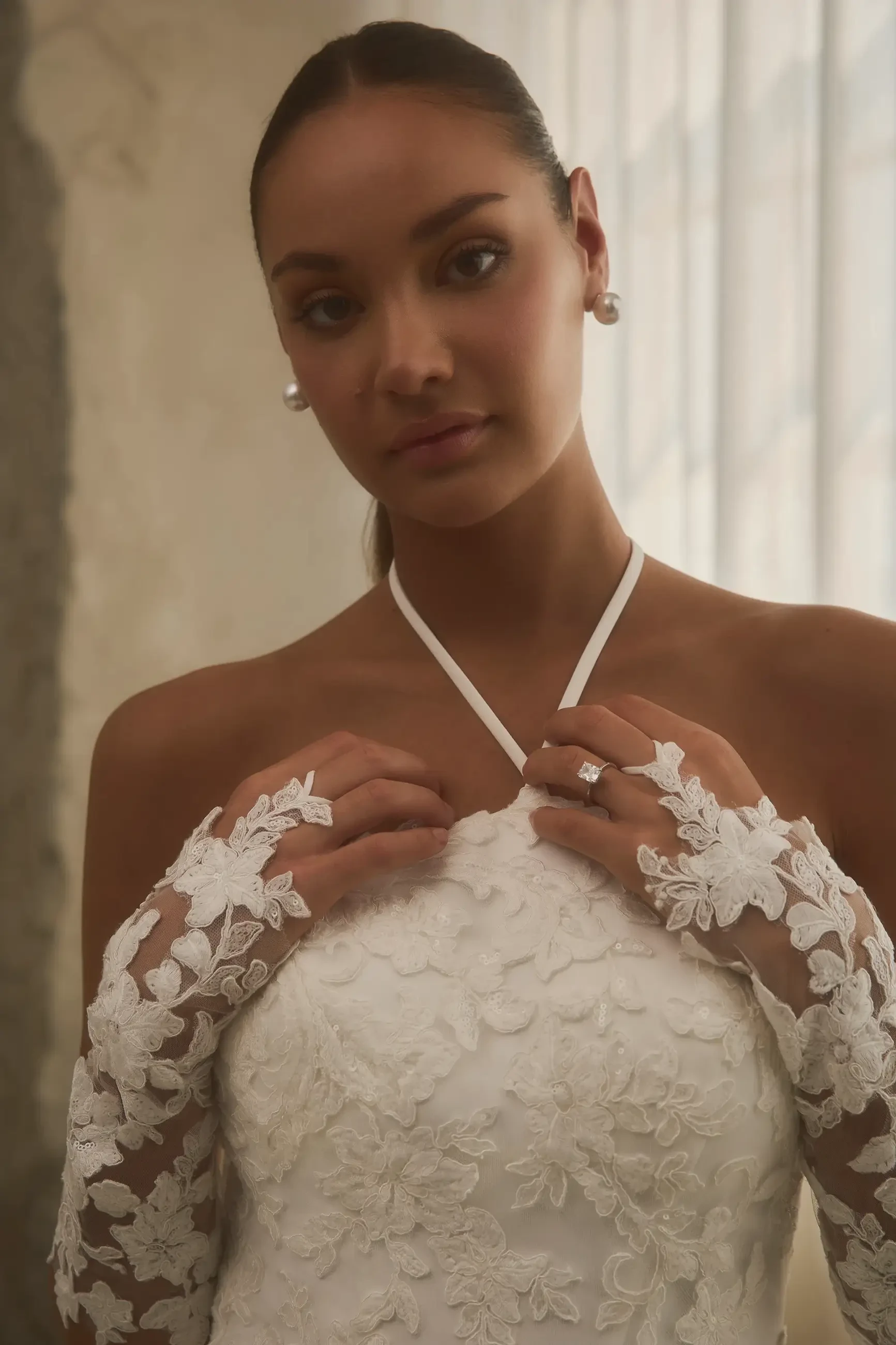 A woman wearing a white wedding dress with intricate lace details and long lace gloves, looking confidently at the camera.