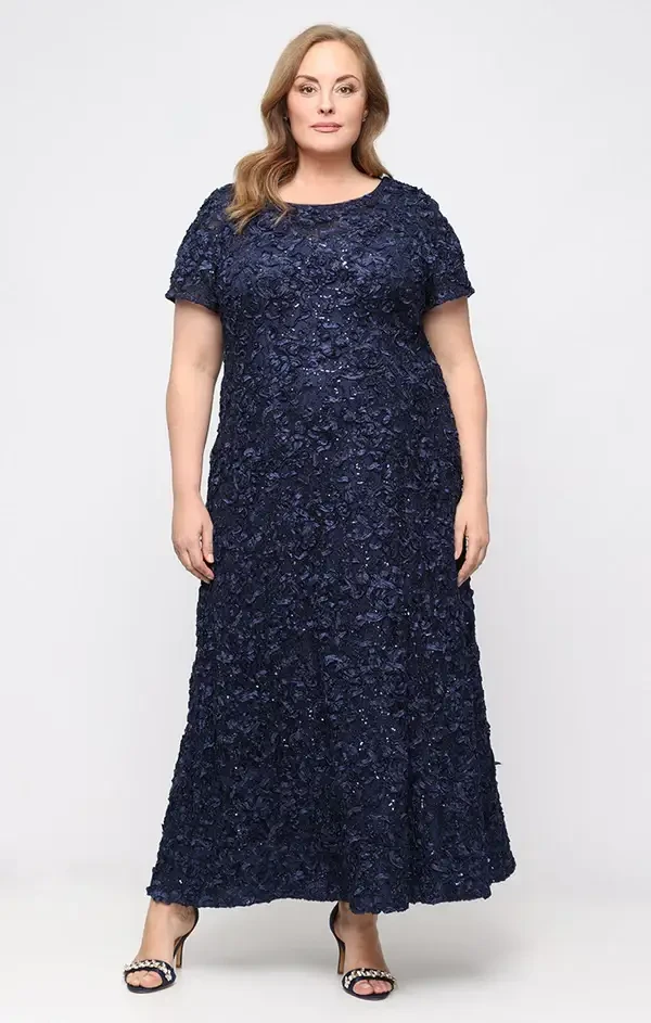 A woman wearing a long, navy blue, floral-embellished dress stands against a plain background. She has long wavy hair and is smiling while posing for the photo.