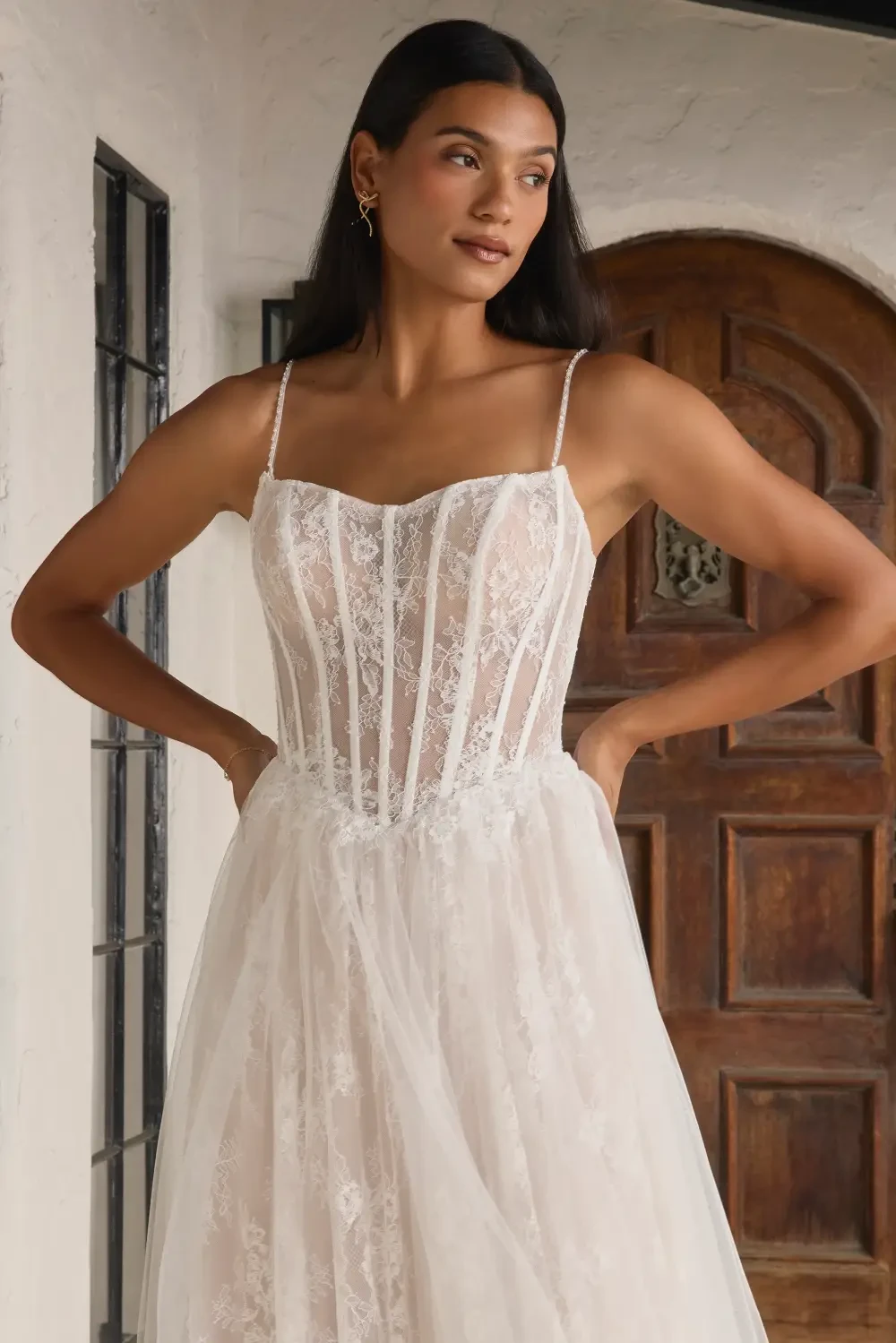 A woman wearing a delicate wedding gown with a fitted bodice and flowing skirt, standing gracefully in front of a wooden door.
