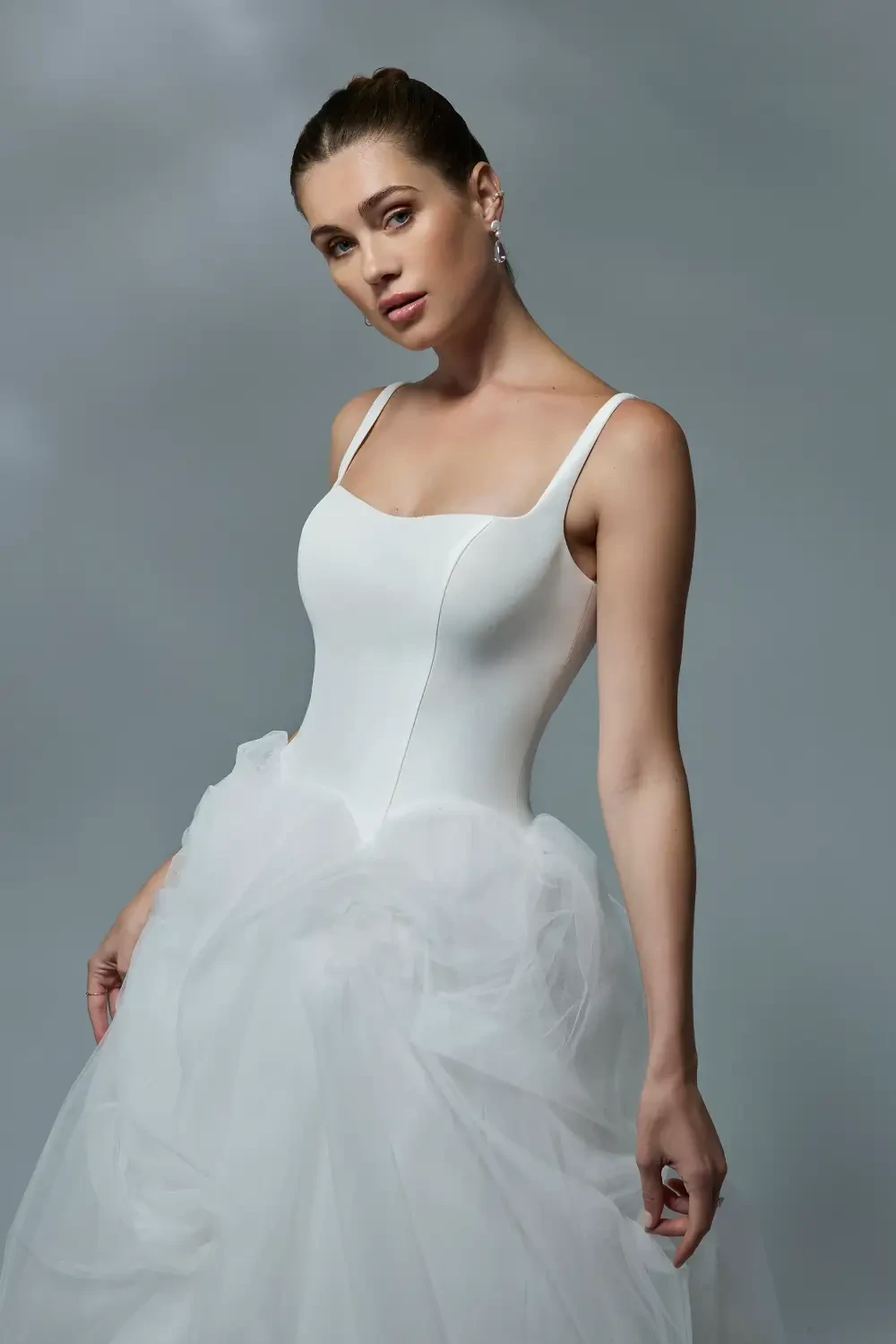 A woman in a sleeveless white dress with a fitted bodice and a flowing tulle skirt, posing against a soft gray background.